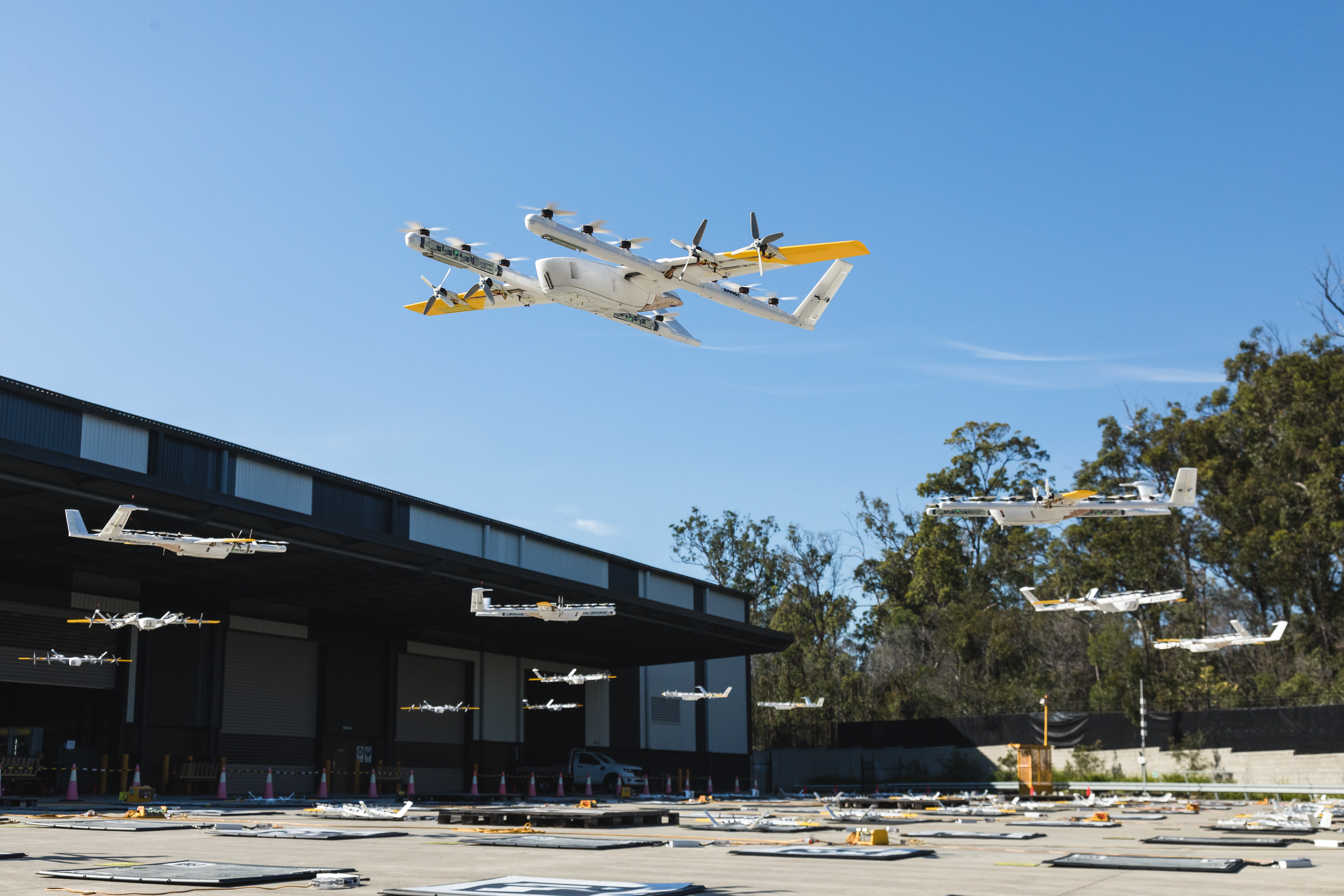 Wing drone hovering over landing pads outside a Wing distribution site. Nine other Wing drones hovering in the background.